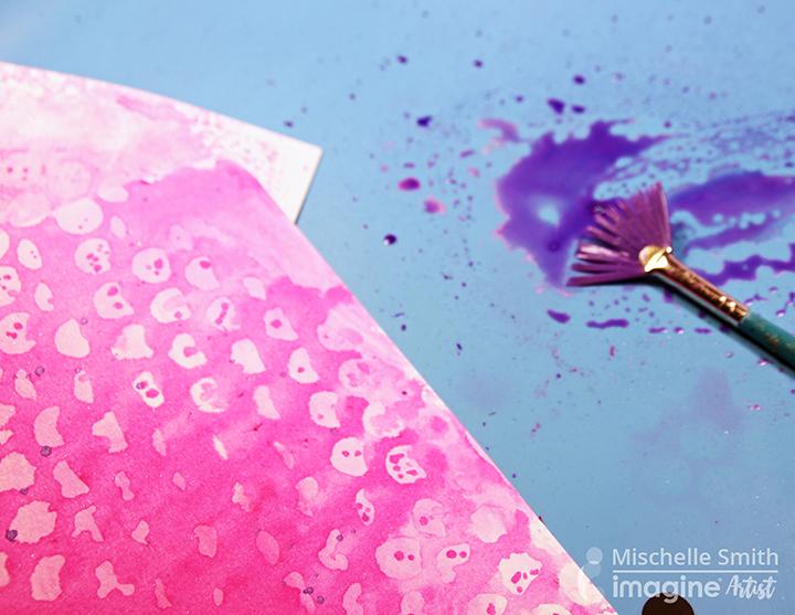 A fan brush with puple Fireworks ink on it next to a pink inked watercolor layout.