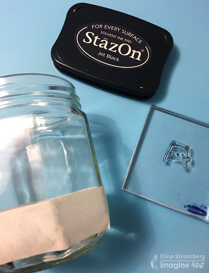 A jar marked with masking tape, a StazOn Jet Black ink pad and an acrylic cat stamp on a stamping block.